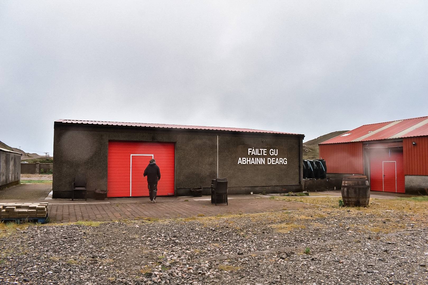Exterior of Abhainn Dearg Distillery: the dark visitor-centre building with a red roller door and the painted Gaelic welcome sign ‘FÀILTE GU ABHAINN DEARG,’ beside a red-roofed outbuilding.
