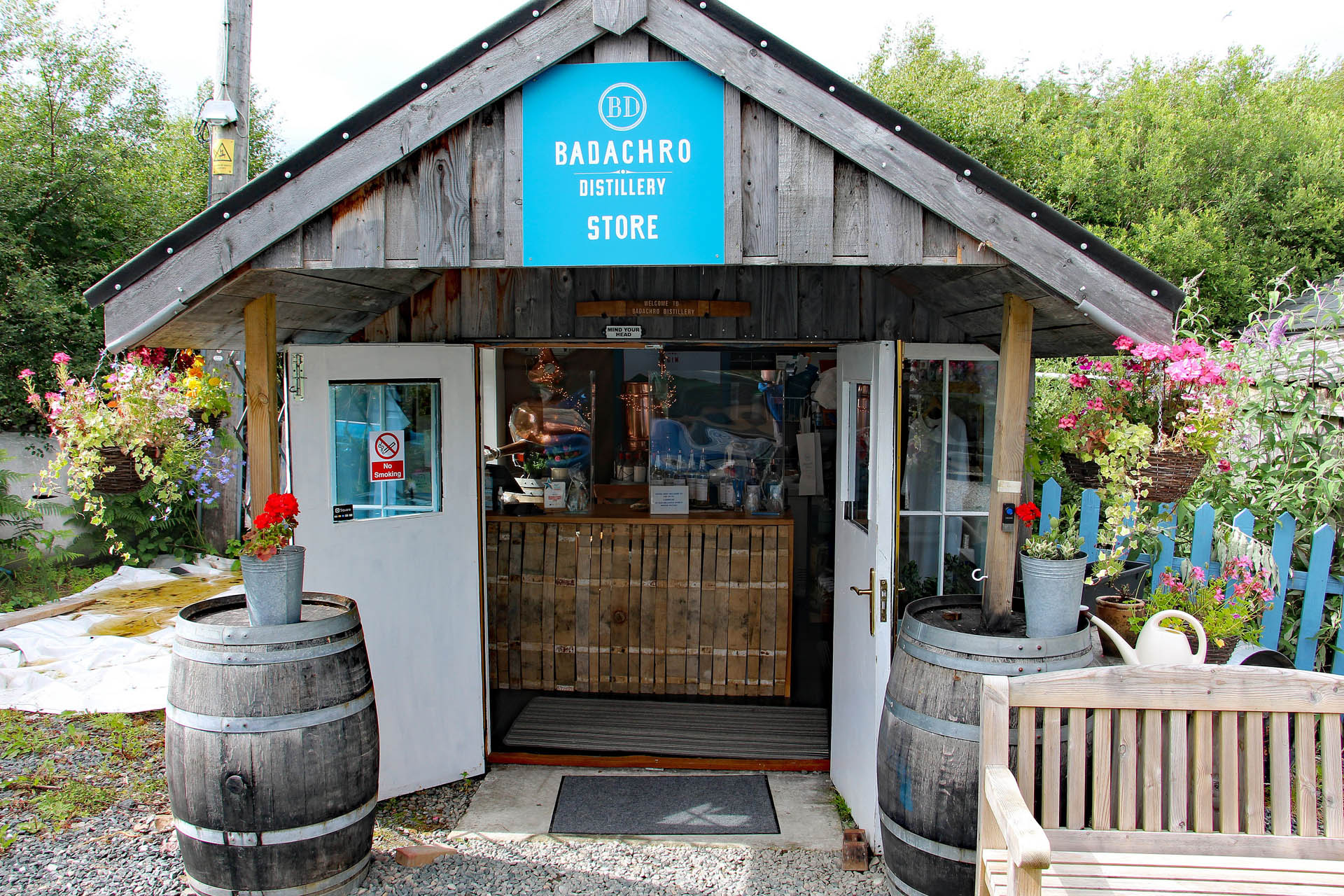 Front exterior of the Badachro Distillery Store: a timber-gabled shop with a blue BADACHRO DISTILLERY STORE sign, open doors, barrel planters, and flowers.