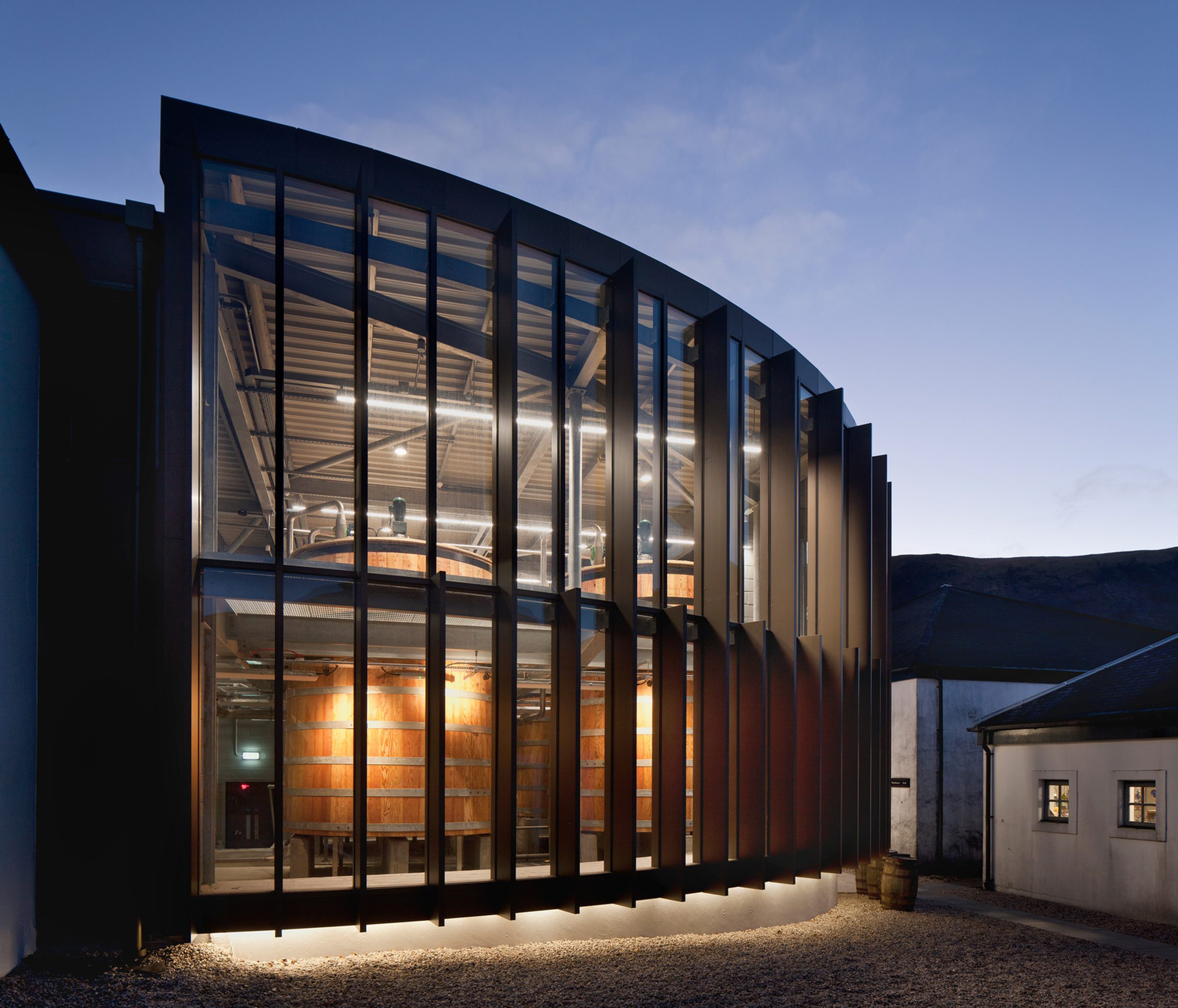 Exterior of Lochranza Distillery’s glazed washback extension at dusk, with timber washbacks visible through the windows and the older white distillery building at right.