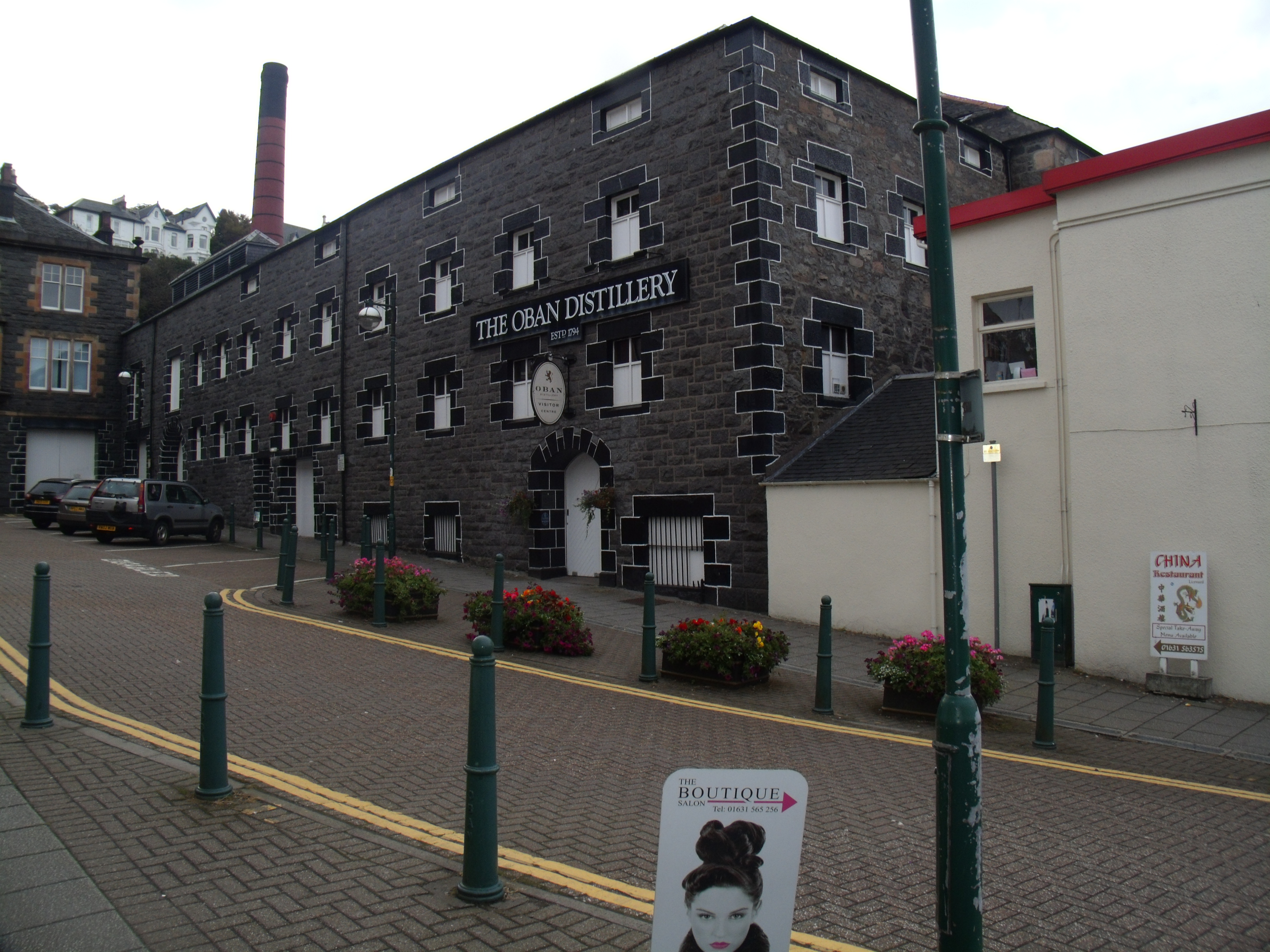 Street-level exterior view of Oban Distillery: a dark stone building with white-trimmed windows, the THE OBAN DISTILLERY sign, and its red-banded chimney.