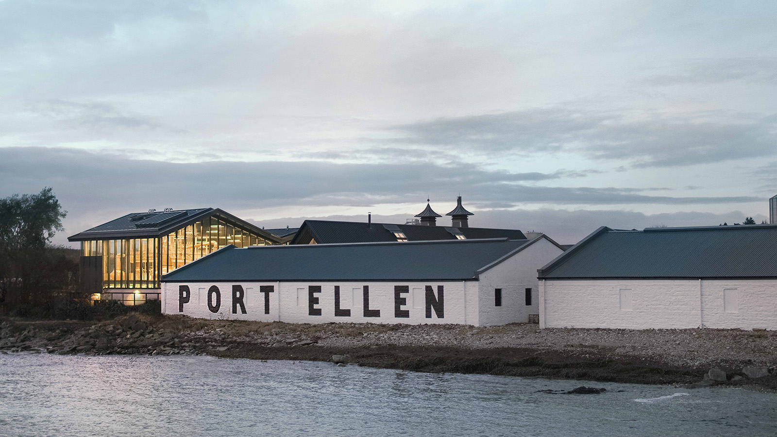 Waterfront exterior of Port Ellen Distillery, with the white PORT ELLEN warehouse in front of the illuminated glass still house.