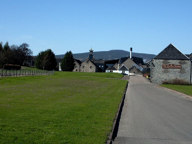 Exterior view of The Balvenie distillery complex in Dufftown, with stone buildings, a tall chimney and a sign reading "The Balvenie Distillery" beside the access road; hills rise behind the site.