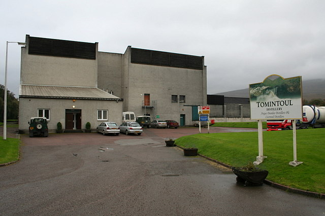 Tomintoul Distillery on the banks of the River Avon, viewed from the entrance drive; the distillery building and TOMINTOUL DISTILLERY sign are visible.